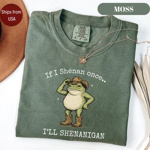 以下が含まれることがあります： カウボーイハットとブーツを履いた漫画のカエルのモスグリーンのTシャツ。「If I Shenan once.. I'll Shenanigan」の文字が下にプリントされています。金のネックレスがシャツの上にかけられています。