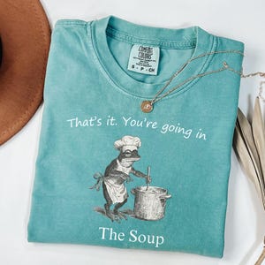 May include: Teal Comfort Colors t-shirt with the text "That's it. You're going in The Soup." A frog wearing a chef's hat and apron is stirring a pot. A gold necklace and a brown hat are also in the image.