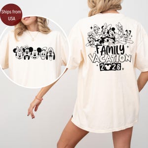 May include: Cream-colored t-shirt featuring a black and white graphic of Disney characters on the front and back. The front displays Mickey, Minnie, Donald, Goofy, and Pluto. The back says "FAMILY VACATION 2026".