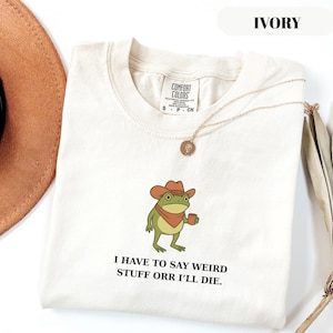 Puede incluir: Camiseta color marfil con un dibujo de una rana con sombrero de vaquero y taza. El texto "I HAVE TO SAY WEIRD STUFF ORR I'LL DIE" está impreso. También se ven un collar dorado y un sombrero marrón.