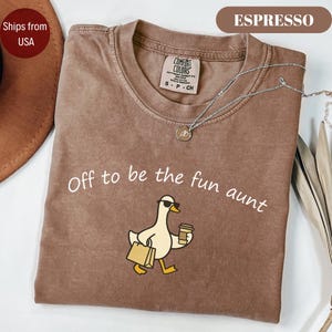 May include: A brown crew neck t-shirt with a graphic print of a white duck wearing sunglasses, carrying a shopping bag and a coffee cup. The text "Off to be the fun aunt" is printed in white above the duck. The shirt has a Comfort Colors label.