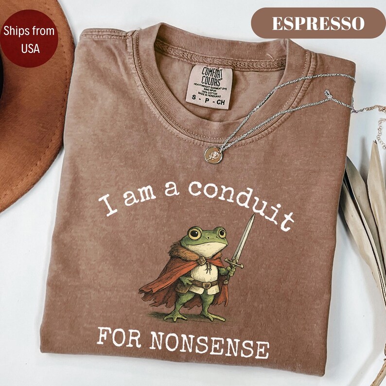 Peut inclure: T-shirt couleur expresso avec l'inscription "I am a conduit FOR NONSENSE" et un dessin de grenouille avec une &eacute;p&eacute;e. Le t-shirt a un col rond et des manches courtes. Un collier argent&eacute; avec un pendentif est sur le t-shirt.