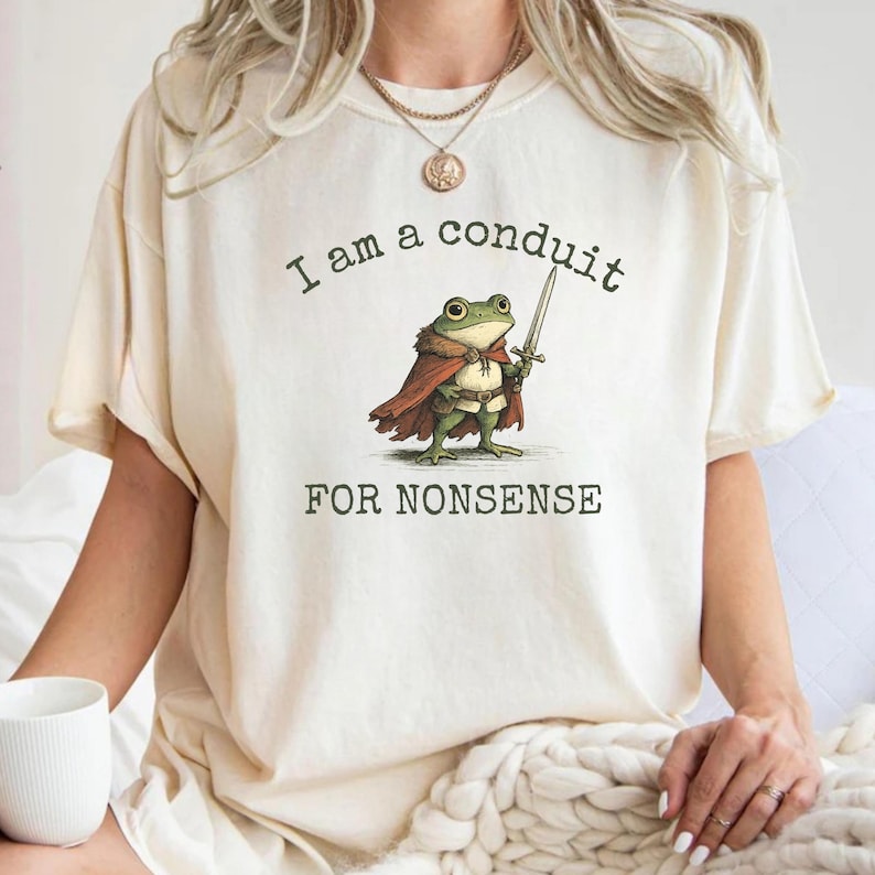 Peut inclure: T-shirt cr&egrave;me avec une illustration fantaisiste d'une grenouille en cape tenant une &eacute;p&eacute;e. Le texte au-dessus de la grenouille dit "I am a conduit", et en dessous, "FOR NONSENSE". Le design est imprim&eacute; sur un tee-shirt doux et d&eacute;contract&eacute;.