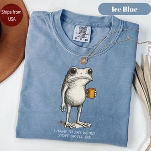 May include: An ice blue t-shirt featuring a graphic of a standing frog holding a small orange mug. Below the frog, the text reads: I HAVE TO SAY WEIRD STUFF OR I'LL DIE. The shirt has a crew neckline and a Comfort Colors brand label at the inner neck.