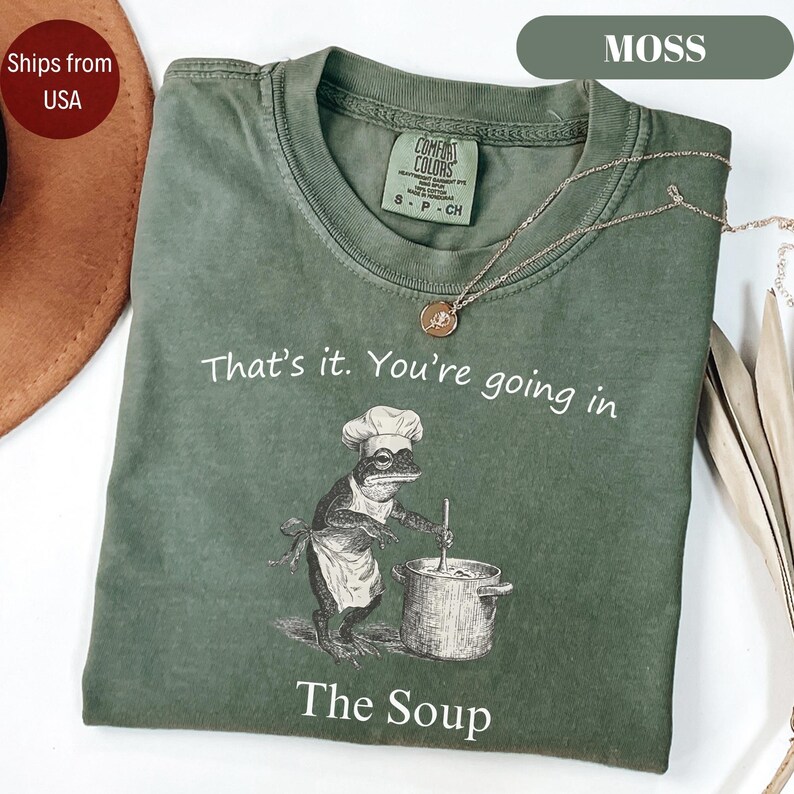 May include: Moss green t-shirt with the text "That's it. You're going in The Soup." A frog chef illustration is printed on the front. The shirt has a crew neck and short sleeves. A gold necklace is draped on the shirt.