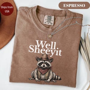 May include: An espresso-coloured t-shirt featuring a graphic of a raccoon wearing a cowboy hat and a vest with a bandana. The text "Well. SHEEYIT" is printed in white above the raccoon. A silver-toned chain necklace rests on the neckline.