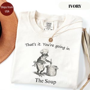 May include: Ivory-colored t-shirt with the text "That's it. You're going in The Soup." A frog chef illustration is featured on the front. A gold necklace with a pendant is draped across the shirt.