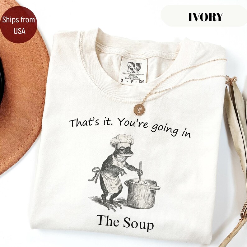 May include: Ivory-colored t-shirt with the text "That's it. You're going in The Soup." A frog chef illustration is featured on the front. A gold necklace with a pendant is draped across the shirt.