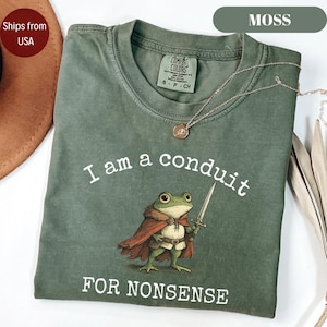 Peut inclure: Un t-shirt vert mousse avec le texte "I am a conduit FOR NONSENSE" et une grenouille de dessin anim&eacute; tenant une &eacute;p&eacute;e. Le t-shirt a un col rond et des manches courtes. Un collier dor&eacute; avec un pendentif est drap&eacute; sur le t-shirt.