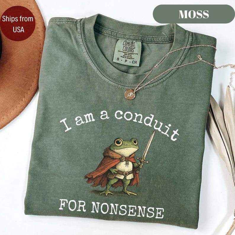 Peut inclure: Un t-shirt vert mousse avec le texte "I am a conduit FOR NONSENSE" et une grenouille de dessin anim&eacute; tenant une &eacute;p&eacute;e. Le t-shirt a un col rond et des manches courtes. Un collier dor&eacute; avec un pendentif est drap&eacute; sur le t-shirt.