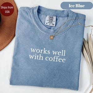 Puede incluir: Una camiseta de manga corta color azul hielo con cuello redondo y acabado teñido en prenda. El frente presenta el texto blanco "works well with coffee" impreso en una fuente sans-serif simple. Un collar dorado con un pequeño colgante circular descansa sobre el cuello.