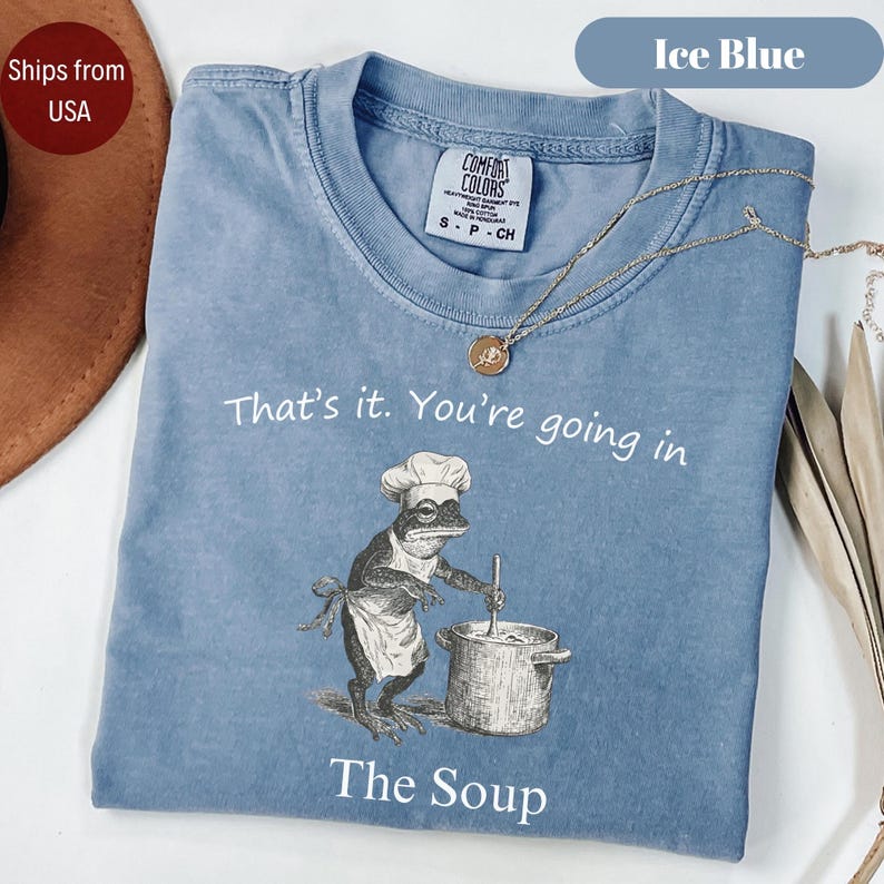 May include: Ice blue Comfort Colors t-shirt with the text "That's it. You're going in The Soup." A frog chef illustration is featured on the front. A gold necklace is draped across the shirt.