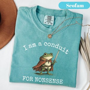 Peut inclure: T-shirt bleu sarcelle Comfort Colors avec l'inscription "I am a conduit for nonsense" et une grenouille de dessin anim&eacute; tenant une &eacute;p&eacute;e. Un collier dor&eacute; avec un pendentif est drap&eacute; sur le t-shirt.