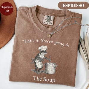 May include: Espresso-colored t-shirt with the text "That's it. You're going in" and "The Soup." The graphic depicts a frog chef stirring a pot. A silver necklace with a pendant is draped on the shirt.