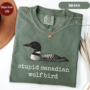 May include: Moss green t-shirt featuring a graphic of a loon with the text "stupid canadian wolf bird". The shirt has a crew neck and short sleeves. A gold necklace with a pendant is draped on the shirt.