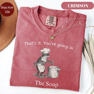 May include: Crimson-colored t-shirt with the text "That's it. You're going in" and "The Soup." The shirt features a frog chef graphic stirring a pot. A silver necklace with a pendant is draped across the shirt.
