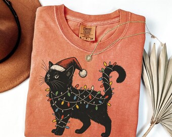 May include: A coral-colored Comfort Colors t-shirt featuring a black cat wearing a Santa hat and wrapped in Christmas lights. The cat is the central graphic, with a gold necklace and the word "YAM" in a peach-colored rectangle.