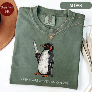 May include: A moss green crew neck t-shirt featuring a graphic of a penguin holding a knife. The text below the penguin reads FLIGHT WAS NEVER AN OPTION. The shirt has a Comfort Colors label at the inner neckline and is paired with a gold necklace.