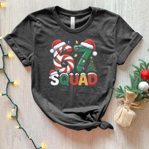 May include: Dark gray t-shirt with a festive Christmas design. The graphic features the numbers "67" in candy cane and green tree designs, with Santa hats and the word "SQUAD" in colorful letters. A holiday-themed tee.