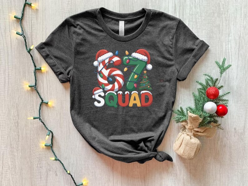 May include: Dark gray t-shirt with a festive Christmas design. The graphic features the numbers "67" in candy cane and green tree designs, with Santa hats and the word "SQUAD" in colorful letters. A holiday-themed tee.