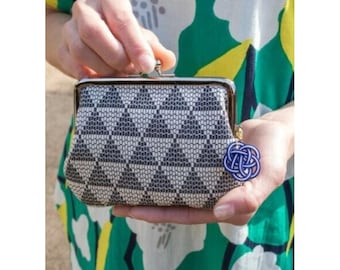 May include: A small, patterned coin purse with a silver clasp. The purse features a geometric design in black and white. A blue and white charm hangs from the side. The purse is held against a background of a green and white floral pattern.