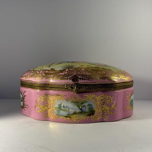 May include: A pink porcelain trinket box with a gold-colored metal clasp and trim. The box features hand-painted landscape scenes and ornate gold embellishments. The lid is curved, and the box has a scalloped edge.
