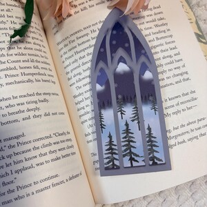 May include: A bookmark shaped like a tall, pointed gothic arch window. The design features a winter forest scene with dark green pine trees, white snow-capped mountains, and a deep blue night sky filled with white snowflakes and stars.