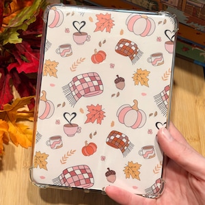 May include: A clear tablet case featuring a repeating pattern of autumn-themed illustrations including pink and orange pumpkins, plaid scarves, coffee mugs with heart-shaped steam, brown acorns, and orange and yellow maple leaves on a cream background.