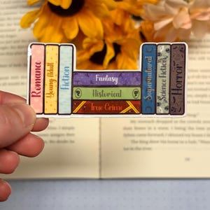 May include: A die-cut sticker featuring a stack of books arranged like a periodic table. The spines are labelled Romance, Young Adult, Fiction, Fantasy, Historical, True Crime, Supernatural, Science Fiction, and Horror in various colours.
