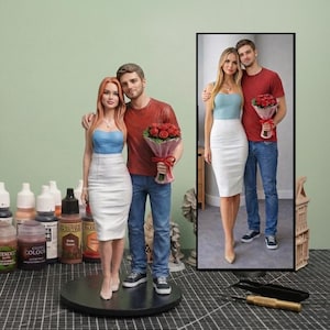 May include: A custom 3D figurine of a couple standing together, modeled after the framed photograph next to it. The female figure wears a blue strapless top and white skirt, while the male figure wears a red t-shirt, blue jeans, and holds a bouquet of red roses.