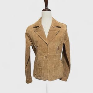 May include: A tan suede blazer with a notched collar, two front pockets, and a single-button closure. The jacket has a fitted waist and long sleeves. The blazer is displayed on a mannequin.