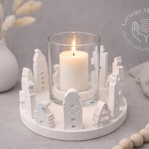 Puede incluir: Un portavelas blanco con un cilindro de vidrio y una vela blanca encendida. El soporte está rodeado de recortes de edificios blancos estilizados. Un jarrón blanco con flores secas está en el fondo. El portavelas tiene unos 15 cm de diámetro.