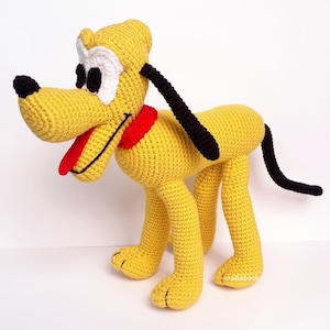 May include: A crocheted plush toy dog in yellow with black ears and tail, red collar, and white eyes. The dog is standing on a white surface.