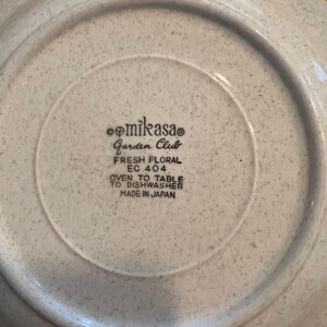 Pu&ograve; includere: Primo piano del retro di un piatto Mikasa Garden Club. Il piatto &egrave; macchiato con un colore grigio chiaro. Il testo sul piatto recita "FRESH FLORAL EC 404 OVEN TO TABLE TO DISHWASHER MADE IN JAPAN."