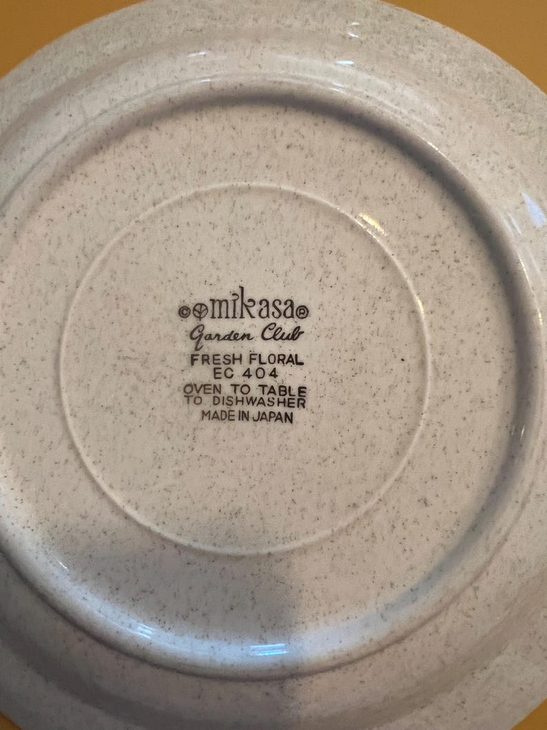 Pu&ograve; includere: Primo piano del retro di un piatto Mikasa Garden Club. Il piatto &egrave; macchiato con un colore grigio chiaro. Il testo sul piatto recita "FRESH FLORAL EC 404 OVEN TO TABLE TO DISHWASHER MADE IN JAPAN."