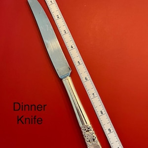 Könnte beinhalten: Ein silbernes Tafelmesser mit einem verzierten Griff, etwa 23 cm lang, wird vor rotem Hintergrund gezeigt. Der Text "Dinner Knife" ist sichtbar. Ein Maßband liegt neben dem Messer.