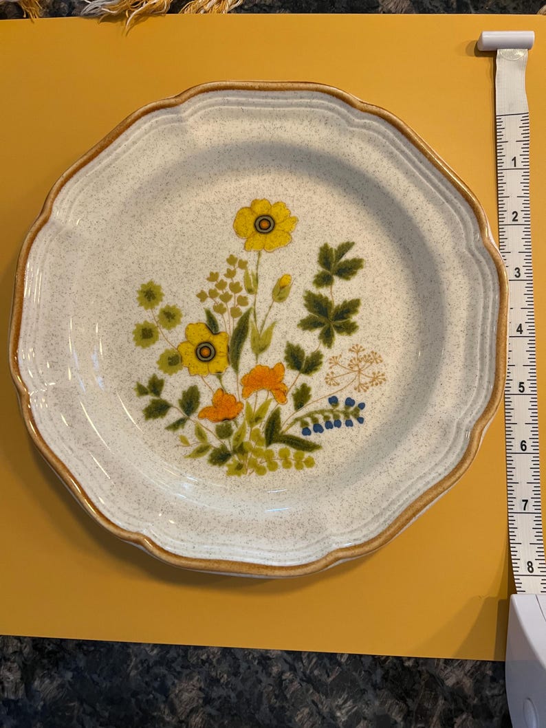Pu&ograve; includere: Un piatto vintage in ceramica con bordo smerlato e design floreale. Il piatto presenta fiori gialli, arancioni e blu con fogliame verde su uno sfondo bianco maculato. Il bordo ha un bordo marrone.