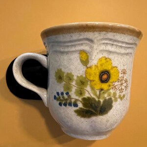 Pu&ograve; includere: Una tazza in ceramica color crema, ornata da un motivo floreale giallo e foglie verdi. Il bordo e il manico sono marrone chiaro. La tazza &egrave; montata su una superficie nera su sfondo giallo.