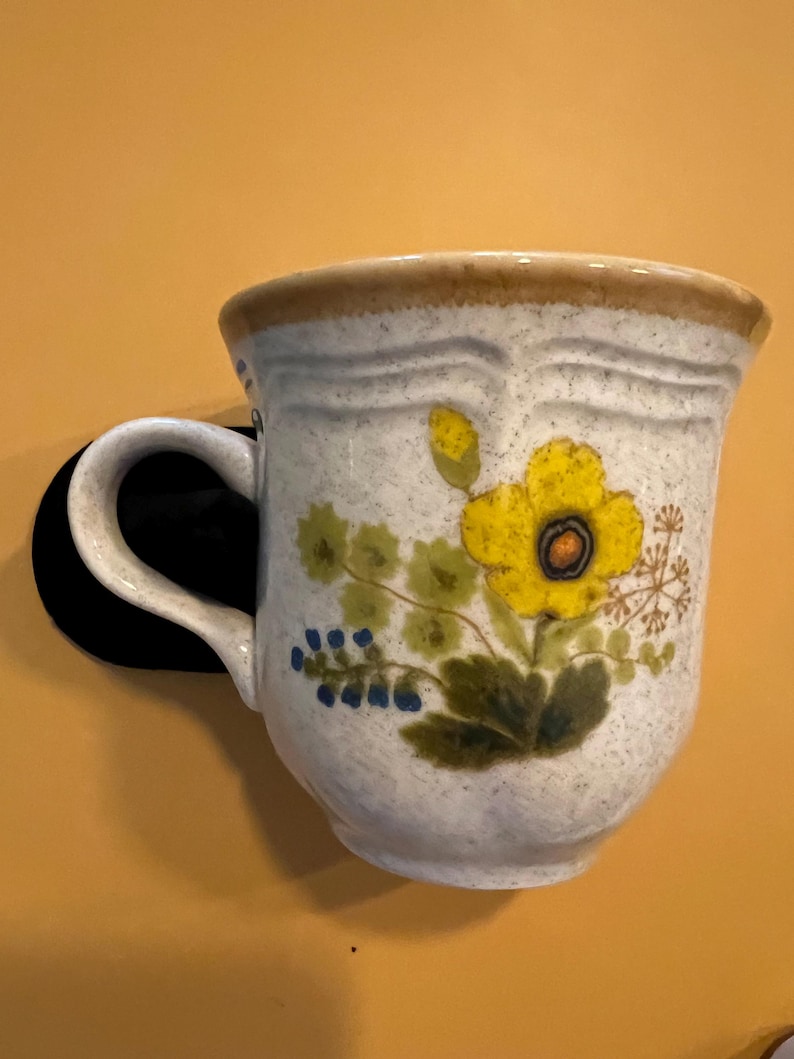 Pu&ograve; includere: Una tazza in ceramica color crema, ornata da un motivo floreale giallo e foglie verdi. Il bordo e il manico sono marrone chiaro. La tazza &egrave; montata su una superficie nera su sfondo giallo.