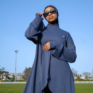 Peut inclure: Un maillot de bain modeste bleu marine avec un col montant et des manches longues. Le maillot de bain présente un design asymétrique et fluide et est associé à un legging noir et un voile noir. Le modèle porte des lunettes de soleil noires.