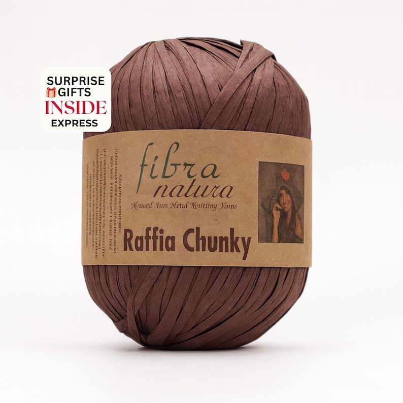 Op de afbeelding: Een bol bruin raffiagaren met een papieren label met de tekst "fibra natura Raffia Chunky". Het label bevat ook een illustratie van een persoon met een hoed. De tekst "SURPRISE GIFTS INSIDE EXPRESS" staat in de linkerbovenhoek.