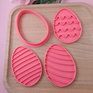 Pode incluir: Quatro cortadores de biscoitos de Páscoa de plástico rosa. Um cortador é de forma oval simples, um tem um design ondulado e dois têm um design listrado.