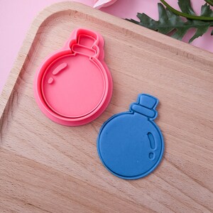 May include: Two cookie cutters, one pink and one blue, shaped like bottles with stoppers. The cutters are on a wooden surface.