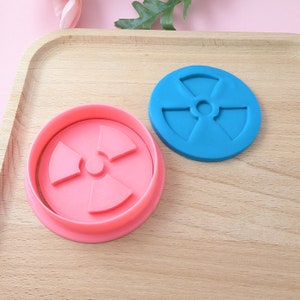 May include: Two plastic cookie cutters in the shape of a radiation symbol. The cutter on the left is pink and the cutter on the right is blue.