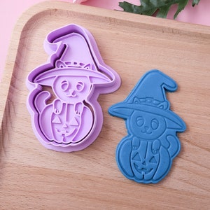Peut inclure: Deux emporte-pièces sur le thème d'Halloween. L'un est violet, l'autre bleu. Les deux représentent un chat portant un chapeau de sorcière et assis sur une citrouille sculptée. Sur un plateau en bois.