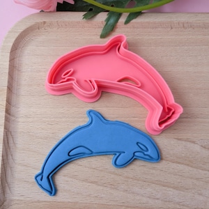 Op de afbeelding: Twee plastic koekjesvormen in de vorm van dolfijnen. De grotere vorm is roze en de kleinere is blauw.