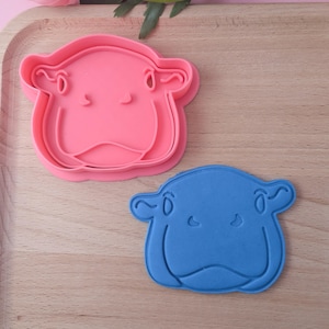 Op de afbeelding: Twee plastic koekjesvormen in de vorm van een nijlpaard gezicht. Eén is roze en de andere is blauw.