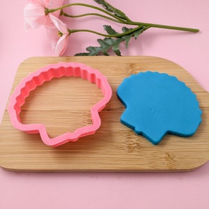 Op de afbeelding: Een roze plastic koekjesvormpje en een blauwe fondant schelp op een houten snijplank.