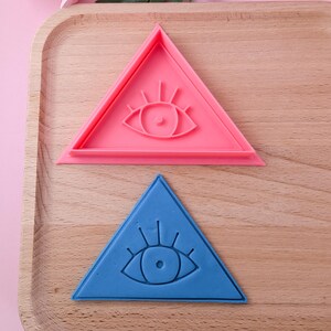 May include: Two triangle-shaped cookie cutters, one pink and one blue, with an eye design in the center.