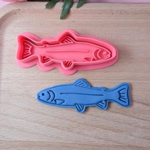 May include: Two fish-shaped cookie cutters, one pink and one blue. The pink cutter is larger and has a more detailed design. The blue cutter is smaller and has a simpler design.